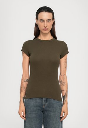 Kvinne med mørkt hår, tatoveringer på armene, iført en tettsittende olivengrønn t-skjorte med korte ermer og blå jeans, stående foran en ensfarget bakgrunn.