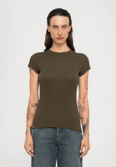 Frau mit dunklem Haar, Tattoos an den Armen, trägt ein enges olivgrünes T-Shirt mit kurzen Ärmeln und blaue Jeans, steht vor einem einfarbigen Hintergrund.