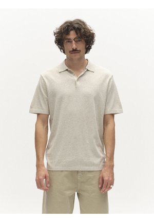 Fiatal férfi göndör hajjal és bajusszal, bézs rövid ujjú galléros ingben és világos színű nadrágban, egyszínű fehér háttér előtt áll.