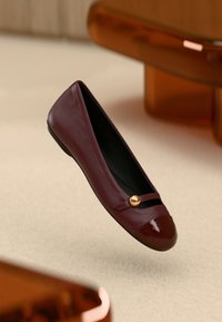 Μπεζ παπούτσι μπαλαρίνας από δέρμα μπορντό με λουστρίνι μύτης, χρυσή λεπτομέρεια και λουράκι στο μπροστινό μέρος. Στερεή μαύρη εσωτερική επένδυση.