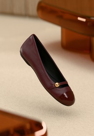 Flacher Schuh aus burgunderrotem Leder mit glänzender Kappe, goldfarbenen Akzenten und einem Riemen über dem Vorderteil. Schwarzes Innenfutter.