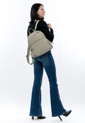 Lichtbeige rugzak met een afgeronde vorm, gewatteerde textuur, verstelbare riemen en een voorzak met rits, gedragen bij donkerblauwe wijde jeans.