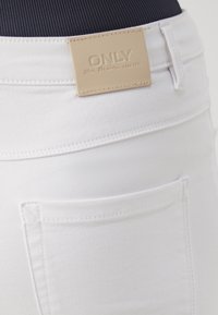 Jeans blancos con un acabado texturizado, que presentan una destacada etiqueta de marca beige en la cinturilla y un bolsillo trasero con costuras sin costura.