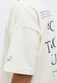 Μπεζ κοντομάνικο πουκάμισο με μεγάλο μαύρο κείμενο στην πλάτη και μικρό γκρι λογότυπο στο μανίκι, φορεμένο από άτομο με μεσαίο τόνο δέρματος.