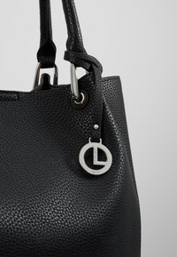 Bolso de cuero negro texturizado con herrajes plateados y una etiqueta con logo circular. Cuenta con dos asas y una abertura espaciosa en la parte superior.