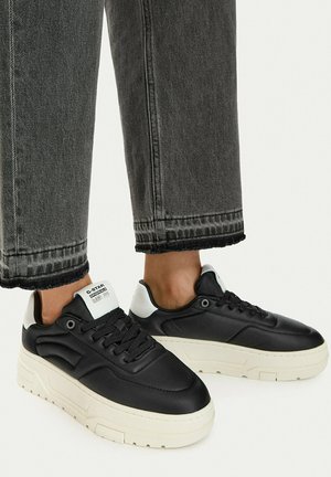 Person iført sort-hvide plateau sneakers kombineret med falmede sorte cropped jeans mod en ensartet baggrund.
