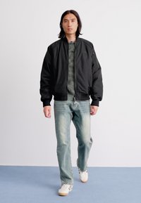 Jeune homme marchant, portant une veste bomber noire, un pull gris, un jean bleu délavé et des baskets blanches sur un sol bleu avec un fond de mur blanc.
