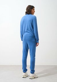 Blå fleece loungewear-set bestående av en långärmad topp och byxor med elastisk midja, med en mjuk textur och figursydd design. Skorna är dolda.