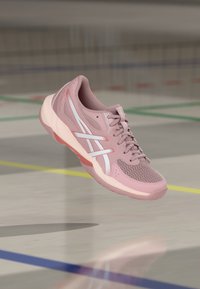 Ροζ αθλητικό παπούτσι με λευκές ρίγες, δεμένα κορδόνια, κρεμασμένο πάνω από το πάτωμα ενός εσωτερικού γηπέδου με πολύχρωμες γραμμές.