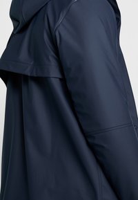 Marine regnjakke med en glat, vandtæt finish. Har hætte, åben krave og let udvidet design. Synlig sidelomme og sømdetaljer.