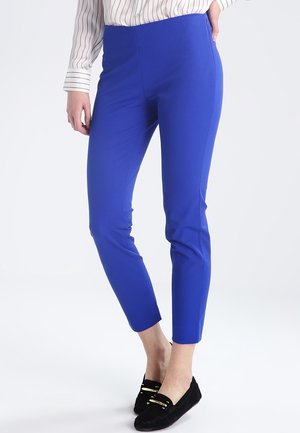 Femme portant un pantalon ajusté bleu vif jusqu'aux chevilles, des chaussures noires sans lacets et un chemisier blanc à rayures verticales noires.