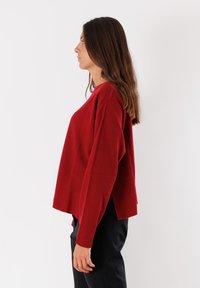 Donna in maglione rosso a coste a maniche lunghe e pantaloni neri in piedi di lato contro uno sfondo bianco.
