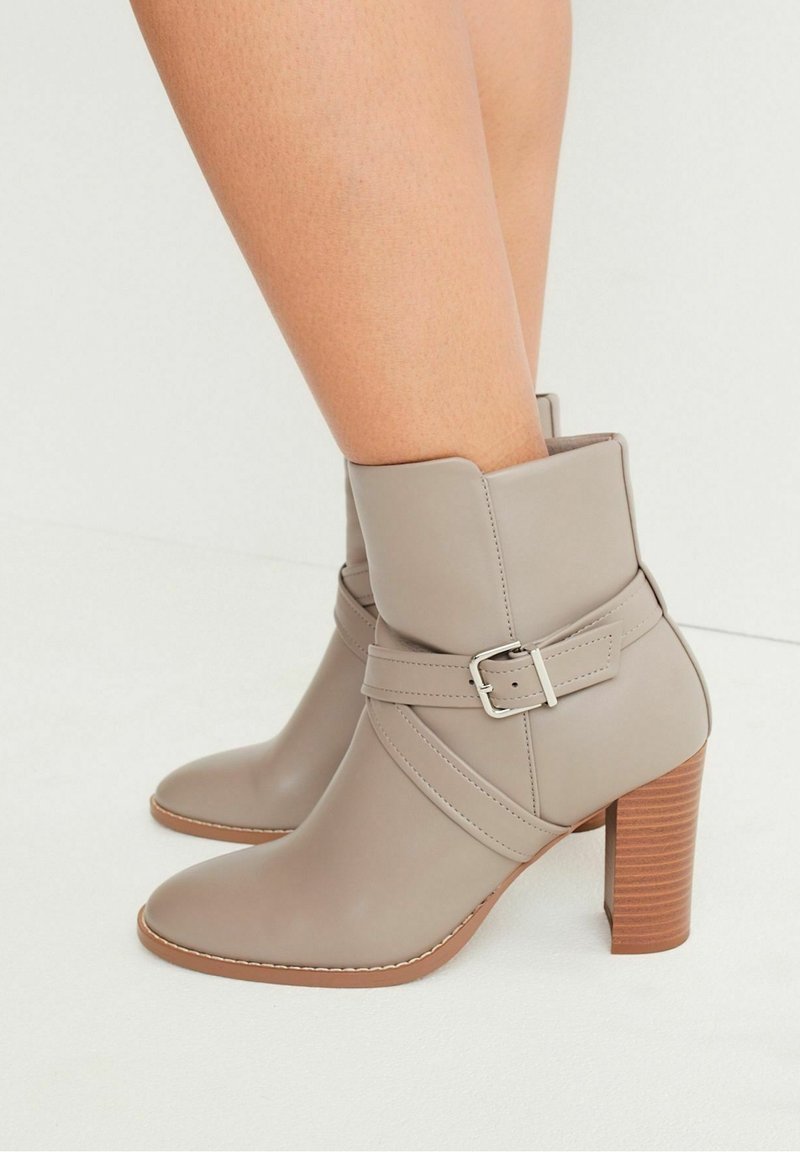 Bottes chevilles en cuir beige avec une sangle à boucle argentée et des talons blocs en bois superposés portées sur des jambes nues.