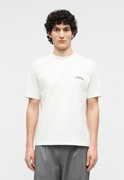 Νεαρός με κατσαρά μαλλιά φοράει απλό λευκό κοντομάνικο μπλουζάκι με μικρό κείμενο "Barbour Feng Chen Wang" στο αριστερό στήθος.