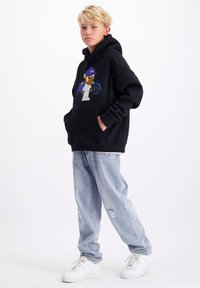 Zwarte hoodie met een cartoonafbeelding aan de voorkant, gecombineerd met lichtblauwe distressed jeans en witte sneakers. Casual streetwear stijl.