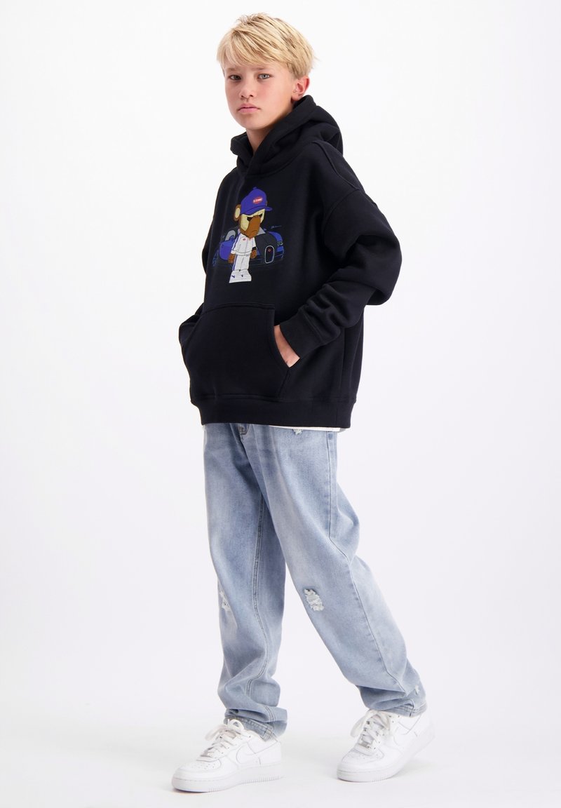 Zwarte hoodie met een cartoonafbeelding aan de voorkant, gecombineerd met lichtblauwe distressed jeans en witte sneakers. Casual streetwear stijl.