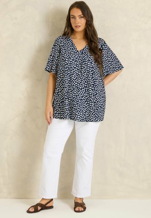 Kvinde iført navyblå kortærmet V-hals bluse med hvide polkaprikker, hvide bukser og brune sandaler, stående foran en beige væg.