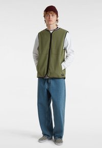 Gilet in pile verde oliva con dettagli neri, chiusura frontale con zip, abbinato a una camicia bianca a maniche lunghe e jeans larghi blu, e scarpe slip-on a scacchi.