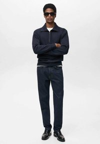 Námornícky zipsový sveter, tmavé denimové nohavice, čierne topánky a slnečné okuliare; obsahuje biele vnútorné tričko a voľný strih. Hladká textúra látky.