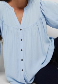 Blouse bleu clair avec un col en V, des détails froncés sous l'empiècement avant et six boutons foncés sur le devant. Tissu doux et texturé.