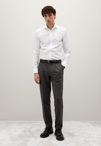 Camicia bianca, pantaloni grigi a fantasia, cintura nera e scarpe stringate nere. Il modello è in piedi contro uno sfondo neutro.