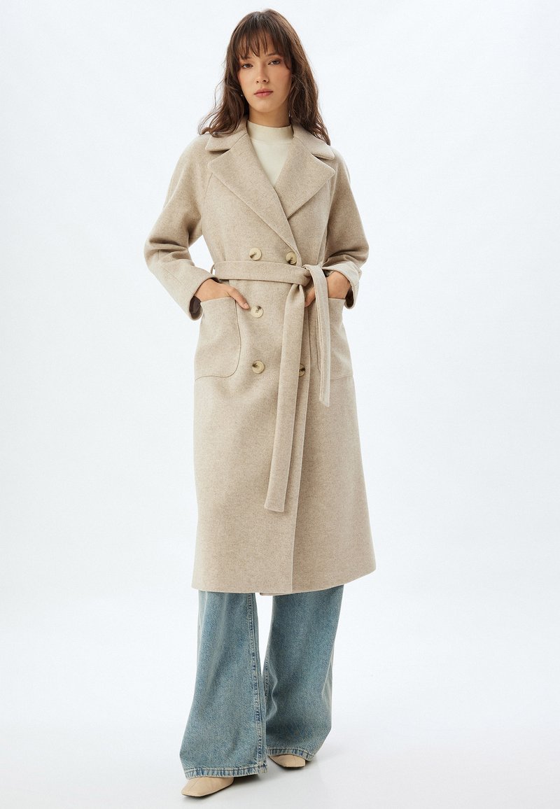 Trench de mezcla de lana beige con un diseño de doble botonadura, grandes botones, bolsillos laterales y un cinturón, combinado con jeans acampanados de color azul claro.