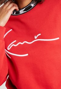 Personne portant un sweat-shirt rouge avec un design brodé blanc de style signature, col à motifs noir et blanc, main touchant le cou.