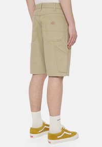 Beige cargo shorts dengan dua saku samping besar dan jahitan yang kontras. Dipakai dengan sneakers suede kuning dan kaus kaki putih hingga pergelangan.