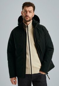Giacca isolante nera con zipper e cappuccio, con sotto un maglione lavorato a maglia di colore crema. I materiali sembrano lisci e resistenti.