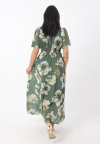 Robe maxi à imprimé floral vert, fabriquée en tissu léger. Détail de taille smockée, manches courtes et poches latérales. Talons visibles en bas.