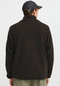 Veste en polaire noire avec un col haut et une surface texturée. Agrémentée d'un patch logo sur l'épaule gauche.