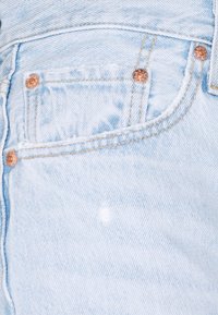 Tejido de denim azul claro con un pequeño bolsillo frontal, herrajes de cobre y detalles de desvanecimiento sutil.