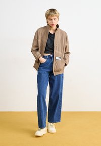 Hellbraune, leichte Jacke mit Kragen, kombiniert mit einem schwarzen Crop-Top und locker sitzenden blauen Jeans. Weiße Sneaker runden das Outfit ab.