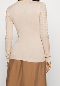 Beige Pullover mit strukturierten Strickärmeln. Figurbetontes Design, runder Halsausschnitt und gerippter Saum. Der glatte Stoff des Körpers kontrastiert mit den gemusterten Ärmeln.