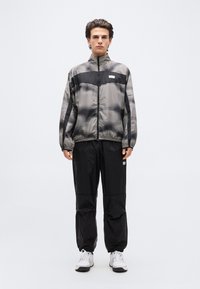 Νεαρός άνδρας που στέκεται φορώντας μαύρο και γκρι ριγέ τζάκετ με φερμουάρ, μαύρο παντελόνι και λευκά αθλητικά παπούτσια, μπροστά σε ένα απλό ανοιχτόχρωμο φόντο.