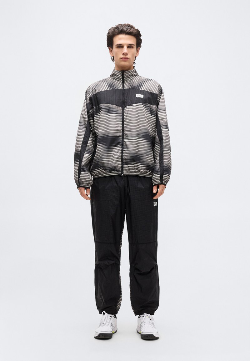 Νεαρός άνδρας που στέκεται φορώντας μαύρο και γκρι ριγέ τζάκετ με φερμουάρ, μαύρο παντελόνι και λευκά αθλητικά παπούτσια, μπροστά σε ένα απλό ανοιχτόχρωμο φόντο.