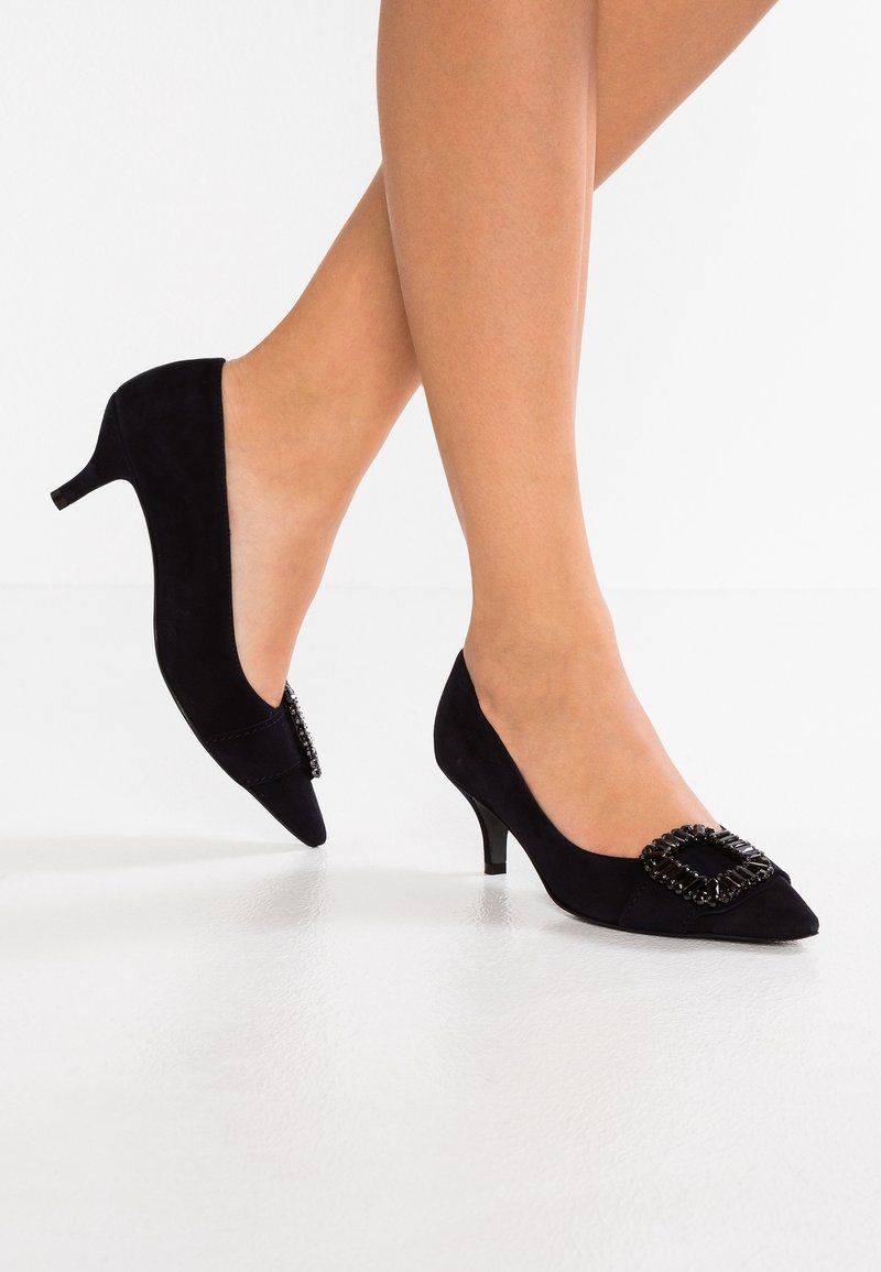 Escarpins en daim noir à talons bas, bouts pointus et boucle noire décorative, portés sur des jambes nues croisées sur fond blanc.