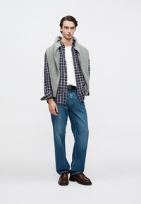 Egy modell szürke pulóvert visel kék-fehér kockás ing fölött, fehér pólóval, kék farmerrel és barna cipővel, egy egyszínű háttér előtt áll.