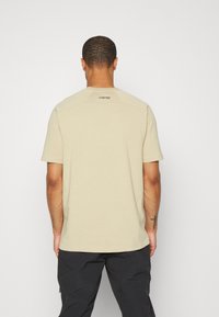 Beige bomulds-t-shirt med korte ærmer, der har rund halsudskæring og et "CORDURA"-logo på den øverste del af ryggen. Enkel, afslappet pasform.