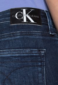 Jeans de denim azul oscuro con una etiqueta negra que presenta el logo "Calvin Klein Jeans" y detalles de costura elegantes en el bolsillo trasero.