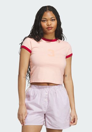 Junge Frau mit langem geflochtenem Haar, trägt ein hellrosa Crop-Top mit rotem Rand und gestreifte Shorts mit Knopfleiste vorne, steht vor einem einfarbigen Hintergrund.