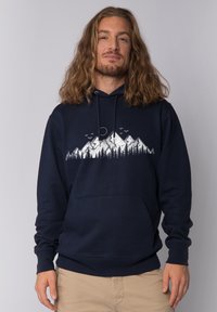 Man met schouderlang haar in een marineblauwe hoodie met een witte afbeelding van een berg en een boom, gecombineerd met beige broek.