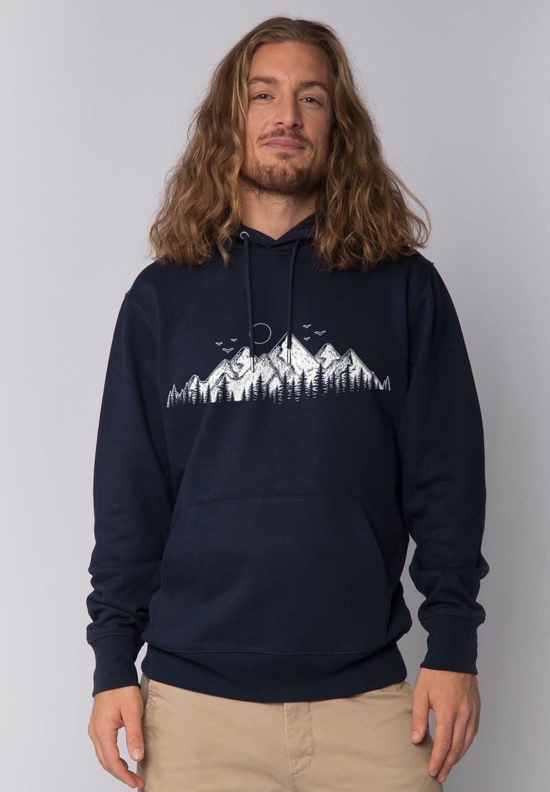 Man met schouderlang haar in een marineblauwe hoodie met een witte afbeelding van een berg en een boom, gecombineerd met beige broek.