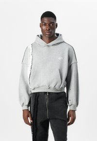 Pelēks īsais hoody ar plīsušiem detalizējumiem vienā pusē, kabata priekšā, kapuce ar aukliņu un brīvi pieguļošām piedurknēm, kombinēts ar melnām biksēm.