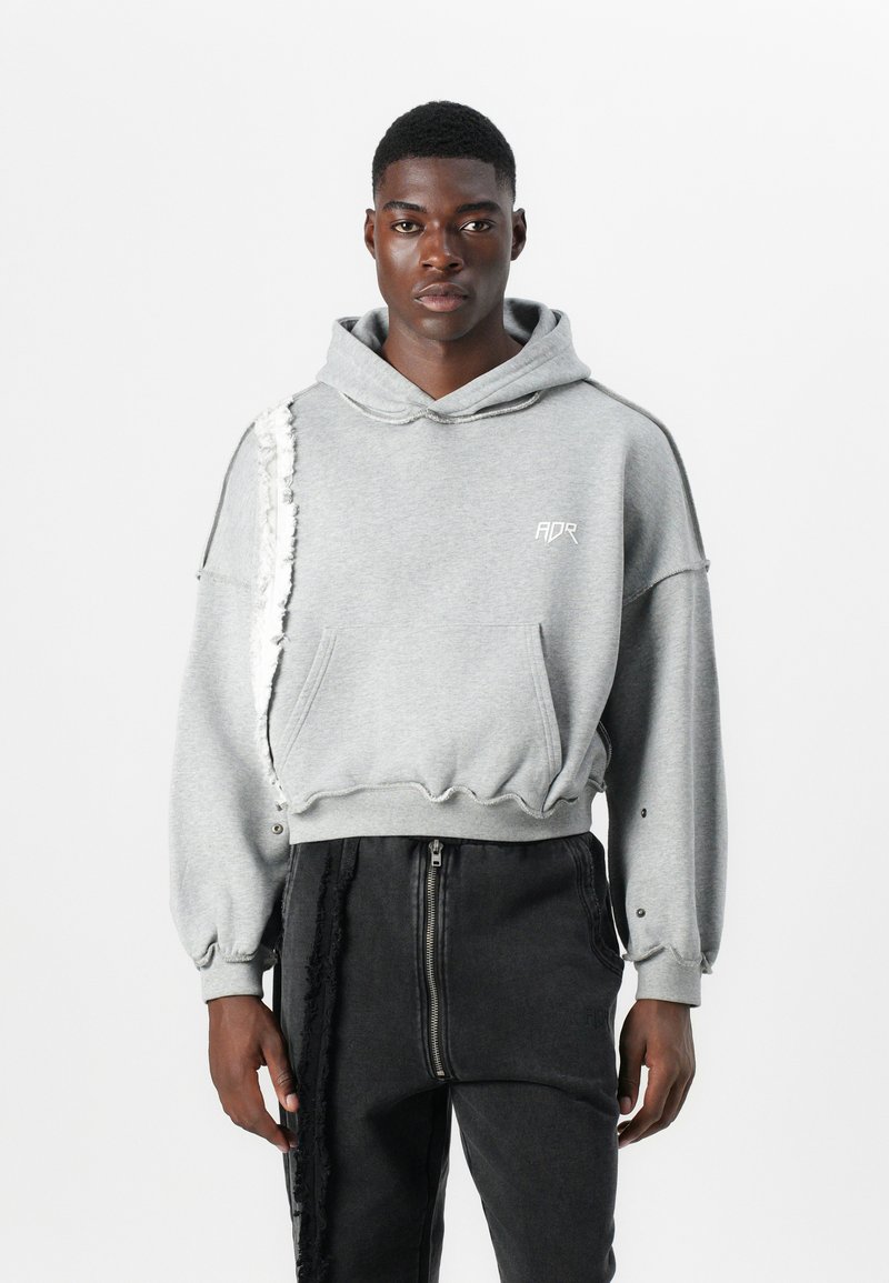 Pelēks īsais hoody ar plīsušiem detalizējumiem vienā pusē, kabata priekšā, kapuce ar aukliņu un brīvi pieguļošām piedurknēm, kombinēts ar melnām biksēm.
