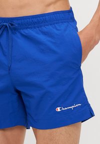 Shorts de bain bleus en matériau léger avec une taille élastique, un cordon de serrage et un logo "Champion" brodé sur l'ourlet.