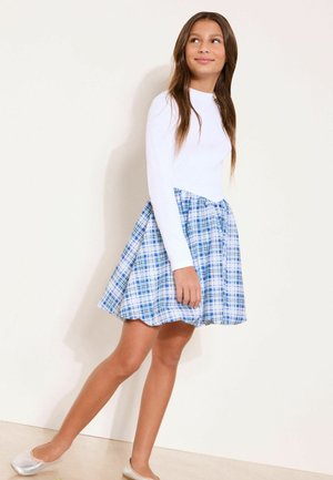 Pige går iført en hvid langærmet top, blå og hvid ternet nederdel samt sølvfarvede balletsko mod en ensartet lys baggrund.