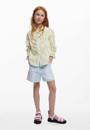 Cămașă cu nasturi în carouri galbene și albe, pantaloni scurți din denim deschis cu margini destrămate și sandale roz cu talpă neagră și curele reglabile.