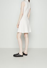 Weißes ärmelloses Kleid mit ausgestellt geschnittenem Saum, gefertigt aus einem strukturierten Stoff, kombiniert mit schwarzen offenen Schuhen mit dicker Sohle.