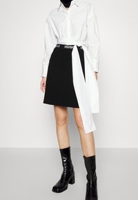 Femme portant une chemise blanche à manches longues avec une ceinture nouée à l'avant, une jupe noire avec la taille HUGO, et des bottes noires à talons mi-mollet.